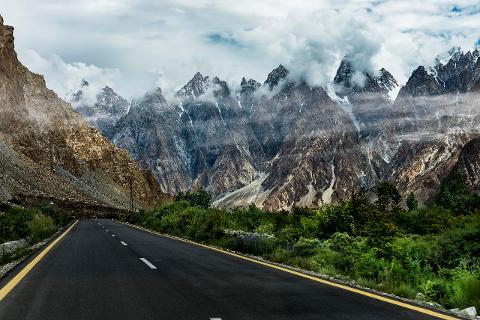 A winding road leads through a mountainous landscape with jagged peaks partially obscured by clouds.
