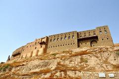 A grand, aged building stands majestically atop a rugged hill, with a clear blue sky as its backdrop.