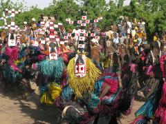A vibrant scene of masked dancers in colorful raffia costumes performing for a crowd of people gathered for a traditional ceremony.