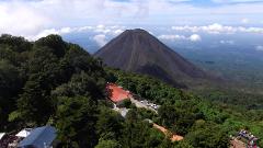A dormant volcano towers over lush green trees, a red-roofed building, parked cars, and a crowd of people in a scenic landscape under a cloudy sky.