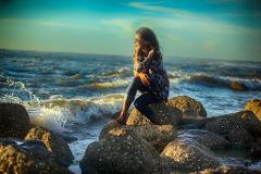 A woman sits pensively on rocks at the beach, gazing at the sea with waves crashing against the rocks under a soft, blue sky.