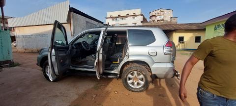 Forfait de voyage à Bangui avec visa à l'arrivée pour la République centrafricaine A silver SUV with its doors open is parked on a dirt lot in front of buildings, with a person in an olive green shirt standing nearby.