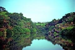 Niger River Odyssey to W National Park and Tapoa 5-Day A tranquil river flows between lush, green cliffs and forests under a clear sky, with the reflections mirroring the scenery on the water's surface.