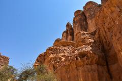 A low-angle shot of a rugged orange rock formation with holes and layered structures under a clear blue sky with bushes at the base.