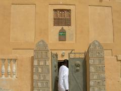 A person in a white robe enters a tan-colored building with a uniquely designed window and door.
