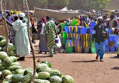 A bustling outdoor market scene with watermelons in the foreground, people browsing, and colorful buckets stacked high, all under a tent-like structure.