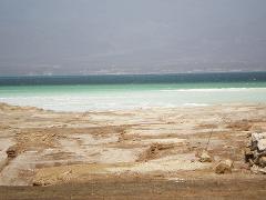 A landscape with a barren, textured terrain leading to a body of water with varying shades of green and blue, under a hazy sky.