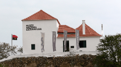 A white building with red tile roof, named "Museu Nacional Escravatura" (National Museum of Slavery), with flags, an Angolan flag, and a stone wall base.