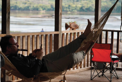 A man wearing glasses and smoking a cigar relaxes in a hammock on a porch overlooking a river, while checking his phone.