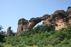 This is a landscape with tall, layered, brown rock formations connected by a natural bridge, set against a clear blue sky and lush green trees.