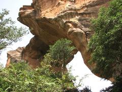 A large sandstone arch is surrounded by green trees, contrasting with the clear sky visible through the archway.