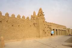 A mosque-like building made of mud bricks stretches across the frame with wooden support beams poking out from the walls.