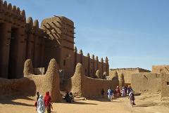 A historic mosque made of mud brick stands tall in a sandy town under a bright blue sky, with people walking along a dirt road in front.