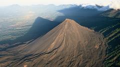 An aerial view captures the imposing, stark beauty of a cone-shaped volcano casting a long shadow over a lush, forested landscape beneath a hazy sky.