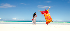 Two Somali women enjoy a beautiful beach day: one in traditional modest attire, the other joyfully expressive in a bright, tie-dye robe with arms outstretched.