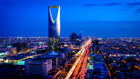 An aerial view captures the glittering Riyadh skyline at night, with the iconic Kingdom Centre tower standing tall amidst a sea of city lights and a highway streaked with car trails.