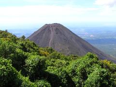 A volcano rises from a lush green forest under a partially cloudy sky.