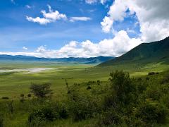 A wide, green valley stretches out beneath a blue sky dotted with fluffy clouds, framed by distant mountains and a lush, green hillside.