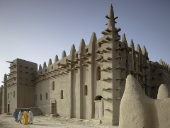 The Great Mosque of Djenné stands prominently, featuring wooden beams and intricate mud construction, with a group of people in colorful robes in front.
