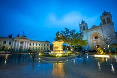 A wide, nighttime shot showcases a city plaza with a statue, cathedral, and government building, all illuminated and reflected in the wet pavement.