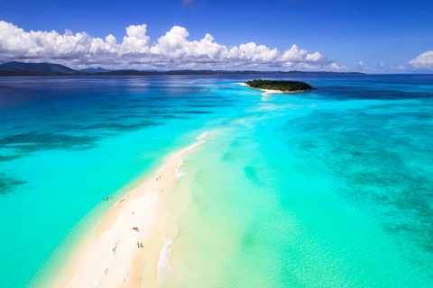 Aerial view of a long, narrow white sandbar leading to a small, forested island, surrounded by turquoise waters under a sunny, cloudy sky.