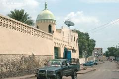 A street scene with a mosque-like building with a dome, a truck in the foreground, and people walking in the background.