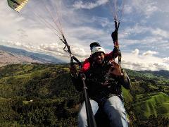 A man with a helmet is tandem paragliding over a lush green landscape, smiling and giving the "OK" sign while holding a selfie stick.