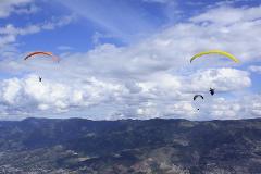 The image shows three paragliders soaring through a partly cloudy sky over a mountainous landscape with urban areas in the valleys.