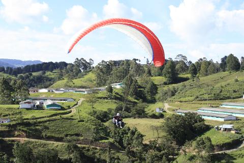 A paraglider soars above a lush green landscape with scattered buildings and hills under a partly cloudy sky.