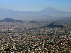 An aerial view of Mexico City sprawling with densely packed buildings, framed by mountains in the background under a hazy sky.