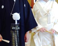 A couple in traditional Japanese wedding attire, the groom in black and the bride in white, hold fans and stand close together.