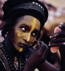 A man with a black turban and intricate yellow face paint applies more makeup using a small mirror at the Gerewol festival.