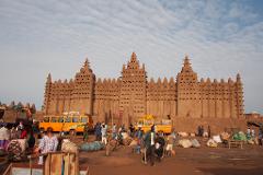 A bustling marketplace in front of the Great Mosque of Djenné, with people, buses, and goods scattered across the dusty ground.