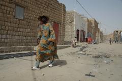 A man in a colorful robe and hat walks down a sandy street in a village, with buildings made of brick and other people in the background.