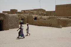 Two girls carry bowls on their heads in an African village with mud-brick buildings on a bright, sunny day.