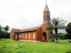 An orange church with a tall steeple stands on a grassy field, surrounded by palm trees under a cloudy sky.