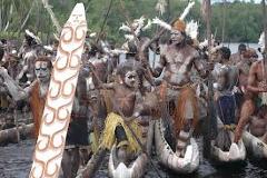 A group of men in traditional attire, including body paint, feathered headdresses, and grass skirts, are in canoes on a river, possibly participating in a festival, holding spears and a large, decorated board.
