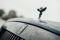 Close-up of a Rolls Royce hood, wet with raindrops, featuring the iconic Spirit of Ecstasy hood ornament and the Rolls Royce emblem.