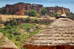 A thatched-roof hut stands in front of a verdant hill with a rocky cliff in the background, under a blue sky.