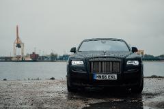 A black Rolls Royce Ghost is parked on a wet, gravelly surface near a body of water, with a crane and industrial buildings visible in the background under an overcast sky.