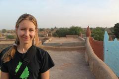A woman in a black t-shirt and camera strap smiles on a rooftop in the Sahel, with traditional mud brick architecture visible in the background.