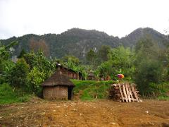 A village scene in Papua New Guinea with a thatched-roof hut, wooden planks, and lush green hills in the background.