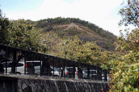 A row of parked cars is partially obscured by a black metal fence and trees, with a hill topped by trees and a trail in the background.