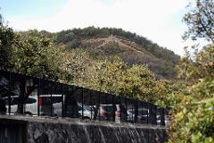 A row of parked cars is partially obscured by a black metal fence and trees, with a hill topped by trees and a trail in the background.