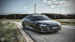 A sleek, black Audi A8 is parked on a rural road, its polished body reflecting the cloudy sky and surrounding greenery.