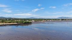 A scenic view of a coastal town under a clear blue sky, featuring a shallow, muddy bay at low tide, with the town's buildings and trees lining the shore and distant mountains in the background.