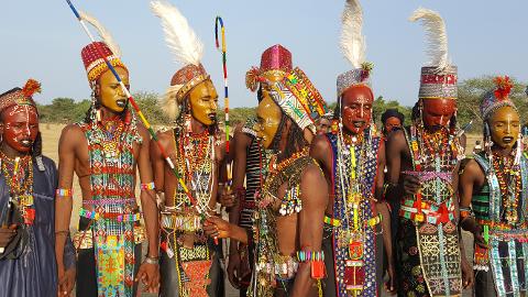 A group of men with painted faces and ornate headdresses and beaded jewelry stand together.