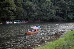 A small boat with passengers is being maneuvered on a river lined with lush green trees and other boats docked on the side.