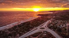 An aerial view captures a stunning sunset over the ocean, meeting a coastline with a town bisected by a highway featuring a roundabout.