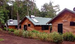 Three wooden cabins with corrugated metal roofs, topped with solar water heaters, sit nestled among lush greenery, creating a serene and eco-friendly lodging scene.